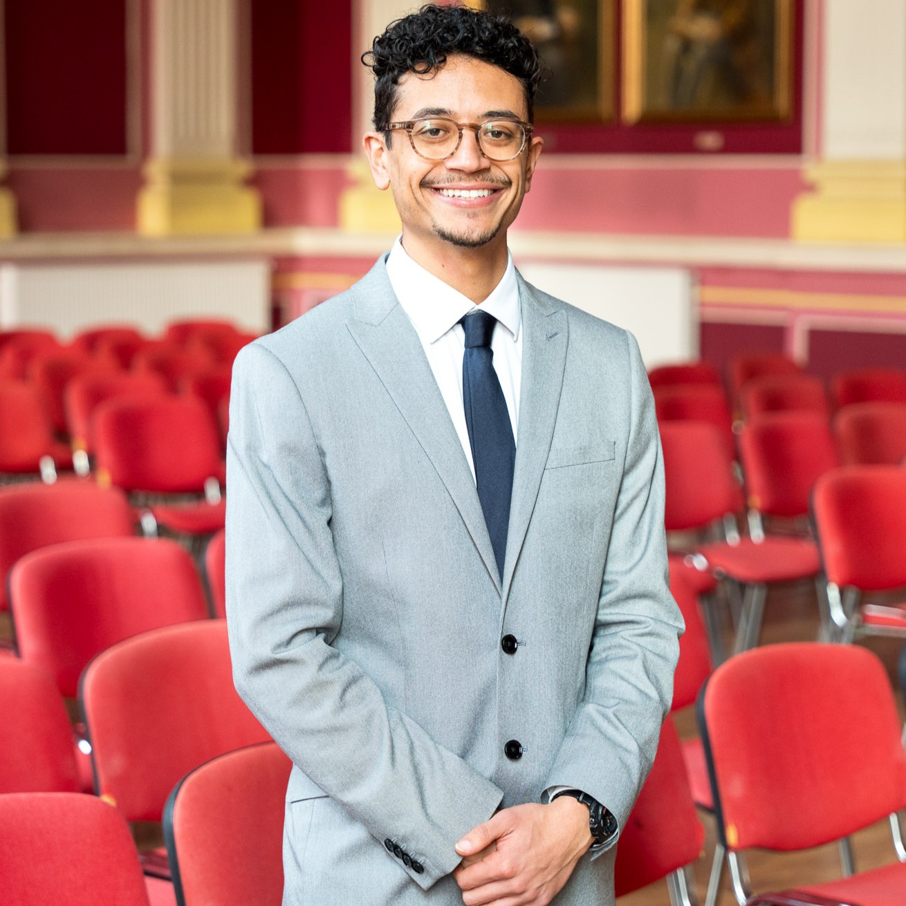 Uśmiechnięty mężczyzna w jasnoszarym garniturze i ciemnym krawacie stoi na tle sali z rzędami czerwonych krzeseł, w eleganckim, klasycznym wnętrzu.