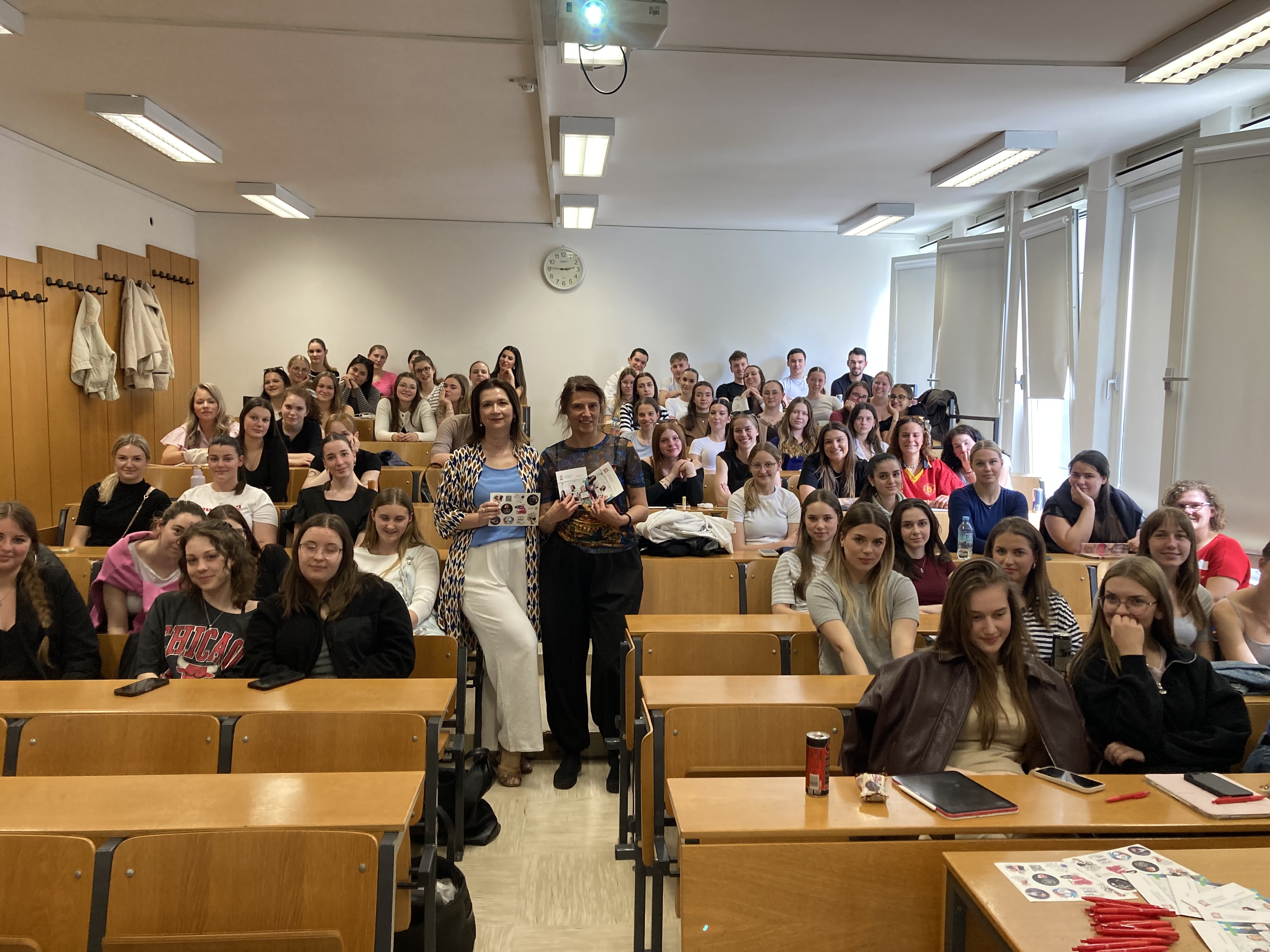 Zdjęcie przedstawia salę wykładową wypełnioną studentami siedzącymi w rzędach drewnianych ławek. Na środku, przed grupą, stoją dwie kobiety trzymające materiały informacyjne. Większość osób patrzy w stronę aparatu i uśmiecha się. Pomieszczenie jest jasne, oświetlone lampami sufitowymi i światłem wpadającym przez duże okna po prawej stronie. Na suficie znajduje się projektor, a na tylnej ścianie wisi zegar. Atmosfera przypomina spotkanie edukacyjne lub wykład.