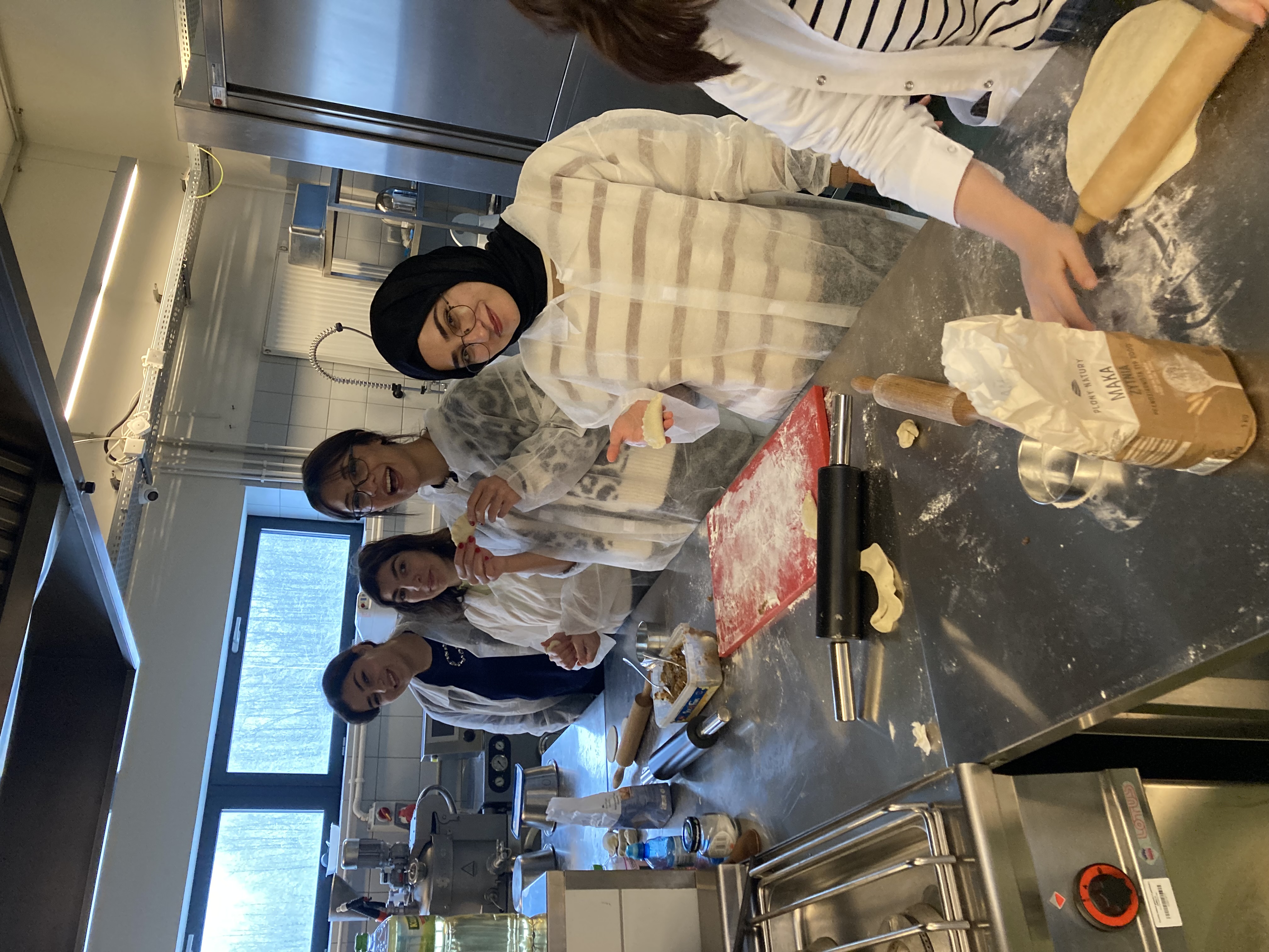 „Grupa czterech młodych kobiet w kuchni szkolnej lub pracowni kulinarnej przygotowuje ciasto. Wszystkie mają na sobie białe ochronne fartuchy. Na blacie leżą wałki do ciasta, mąka i akcesoria do pieczenia. Jedna z kobiet trzyma kawałek ciasta i uśmiecha się do aparatu, pozostałe też wyglądają na zaangażowane i zadowolone. W tle widać sprzęt kuchenny i duże okno z widokiem na zimowy krajobraz.”