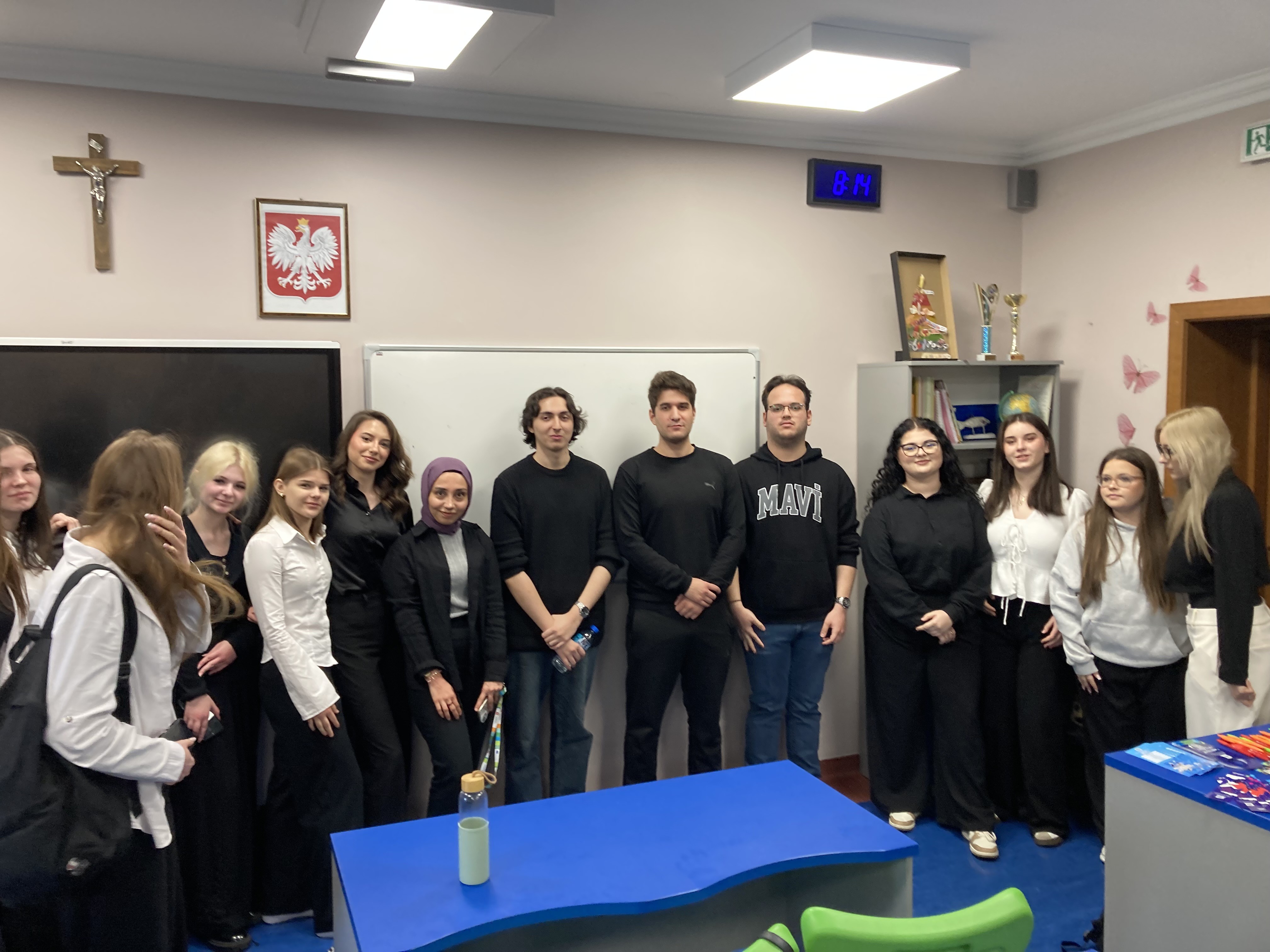 Grupa kilkunastu uczniów stoi w klasie przed tablicą, ustawiona w rzędzie i pozująca do zdjęcia. Większość osób ubrana jest w czarne lub biało-czarne stroje. Nad nimi na ścianie widoczny jest krzyż oraz godło Polski. Po prawej stronie stoi regał z książkami i pucharami. W pomieszczeniu znajduje się także niebieskie biurko z butelką na wodę oraz zielone krzesło. Na ścianie widać zegar elektroniczny wskazujący godzinę 8:14.