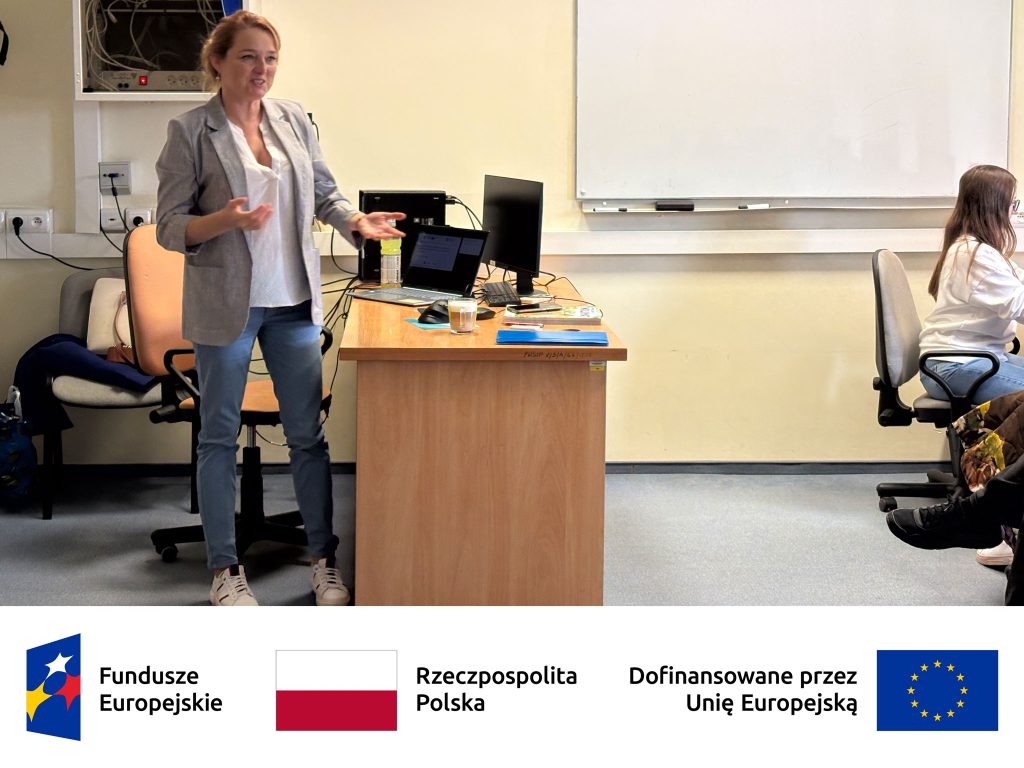 „Prowadząca stoi przy biurku w sali szkoleniowej i gestykuluje podczas prezentacji. Na biurku znajdują się laptopy, dokumenty i napoje, a w tle widać tablicę oraz uczestniczkę siedzącą przy komputerze. Na dole grafiki widoczne są logotypy Funduszy Europejskich, Rzeczypospolitej Polskiej i Unii Europejskiej