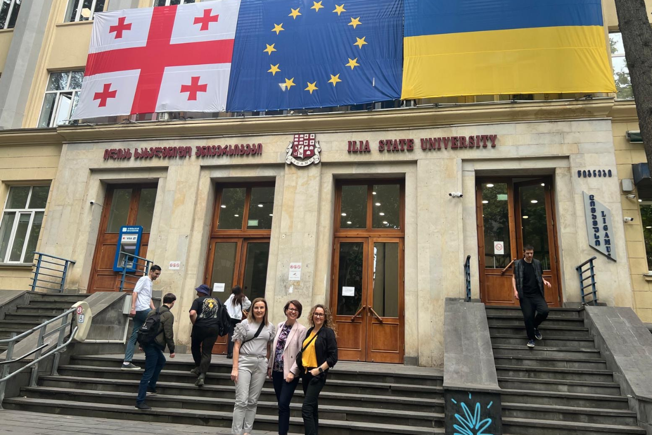 Trzy Panie stojące plecami do budynkuI lia State University. U szczyty budynku znajdują się trzy ogromne flagi, od prawej Gruzji, Unii Europejskiej oraz Ukrainy