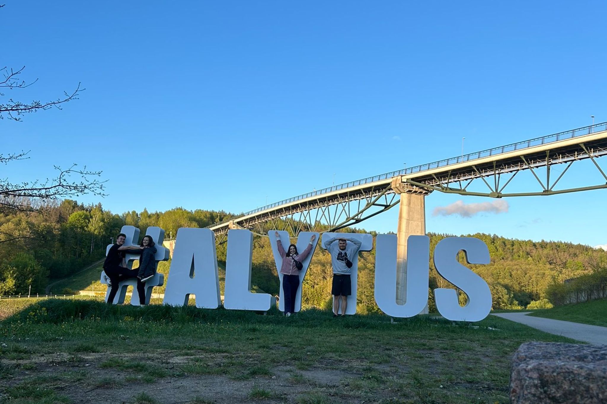 Grupa studentów wpasowuje się w litery napisu ALYTUS na tle mostu