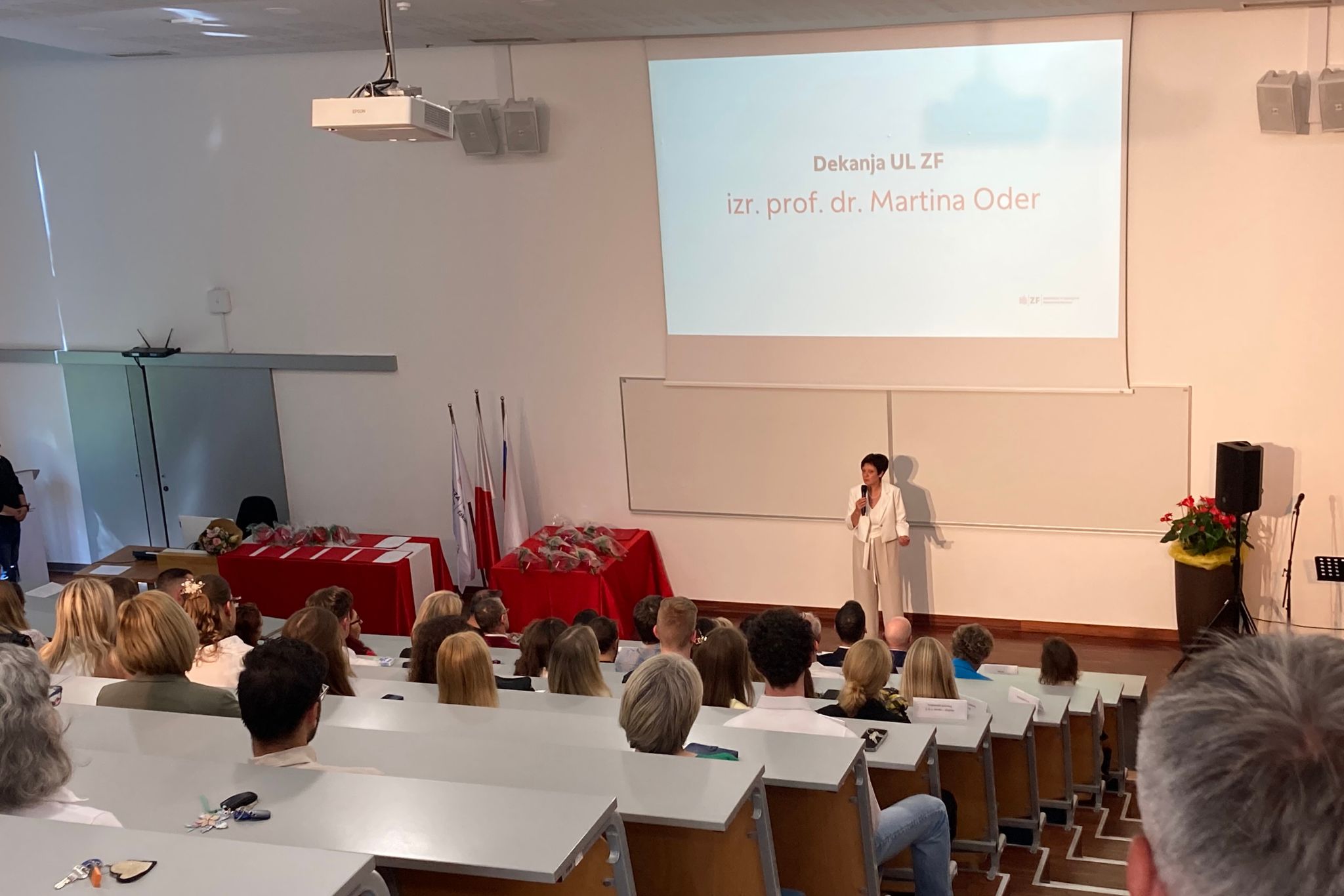 Sala wykładowa. Na dole stoi wykładowca przedstawiający wyświetloną prezentację.  W ławkach na podwyższeniu widać dużą grupę ludzi.