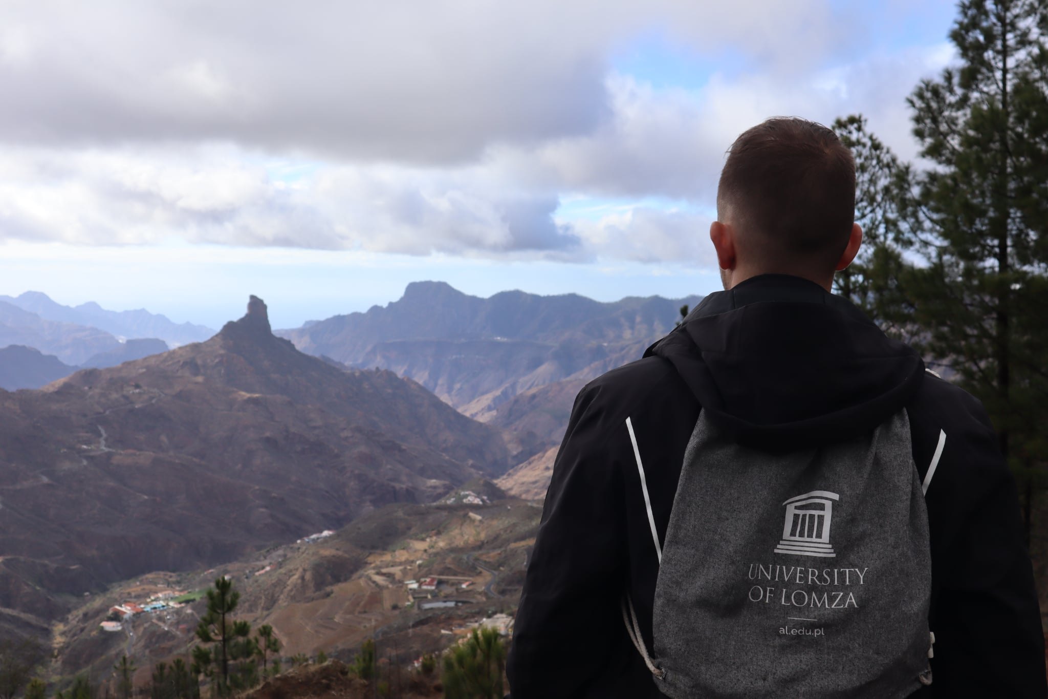 Mężczyzna z plecakiem na plecach na którym widać logo University of Lomza, patrzy w stronę gór