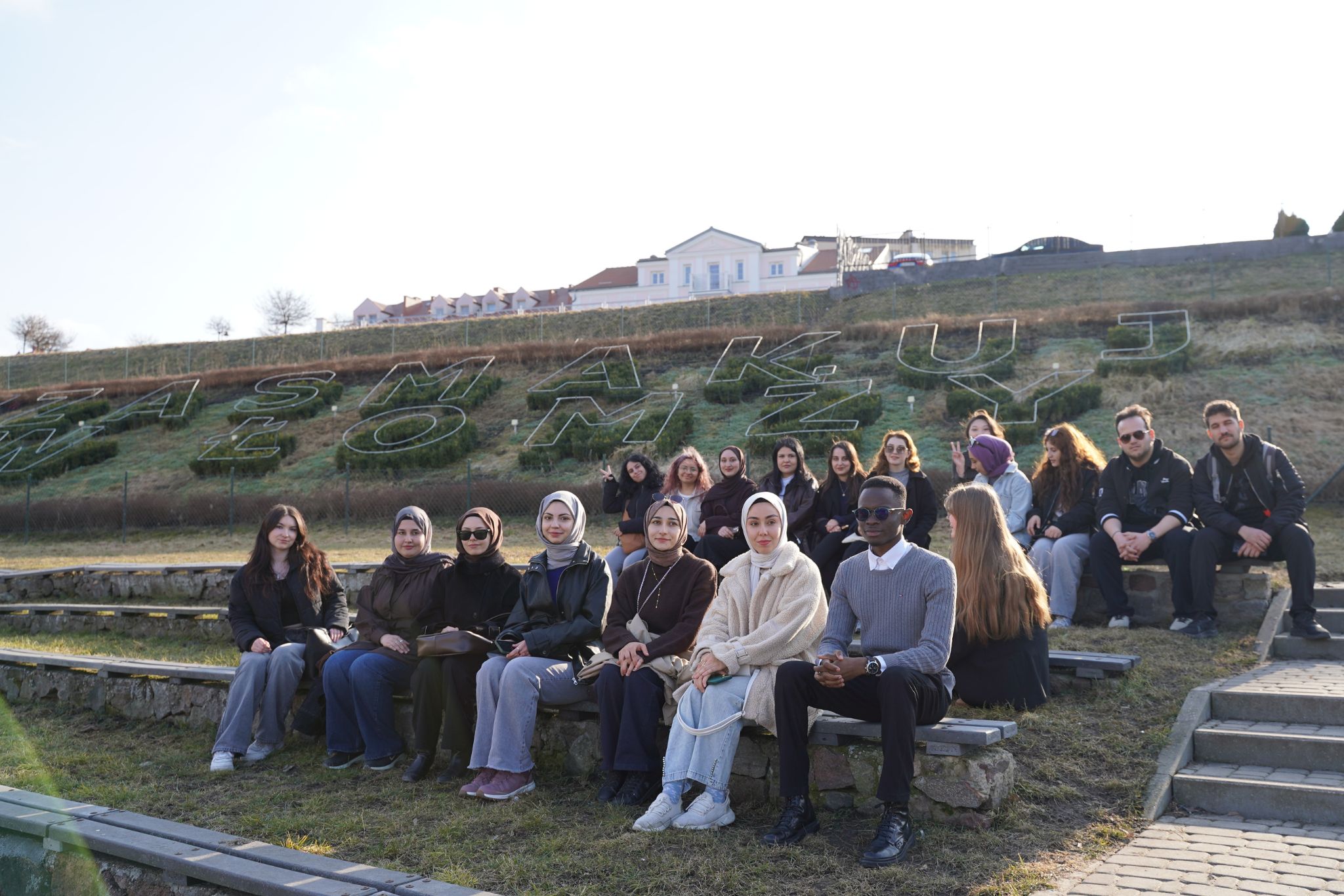 Grupa osób pozuje do zdjęcia siedząc na ławkach, na muszli koncertowej. W tle widać napis Zasmakuj w Łomży