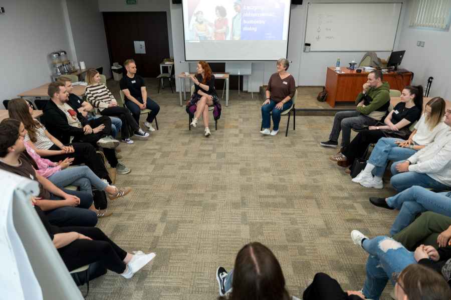 grupa studentów siedzi na krzesłach w okręgu w sali zajęciowej, w tle ekran rzutnika