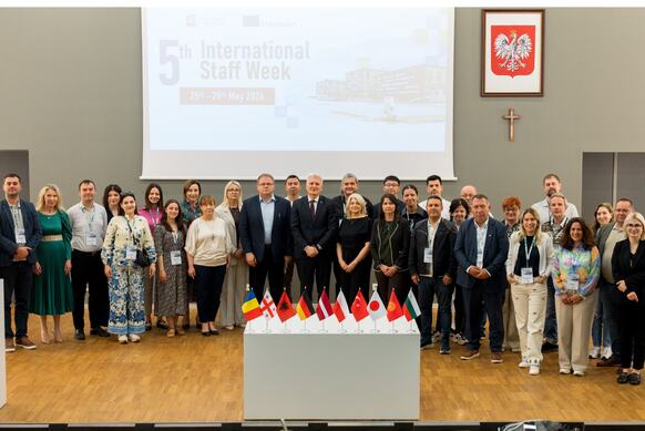 A group of people are posing for a photo. In front of them is a table with 10 flags representing different countries; behind them is a slide displaying the name of the event