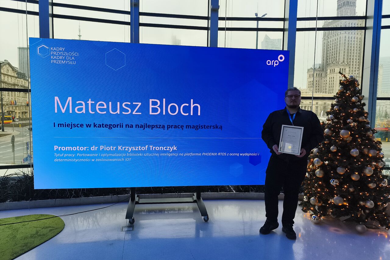 A person stands next to a large blue screen displaying the text: “Mateusz Bloch – First Place in the category for Best Master’s Thesis. Supervisor: Dr Piotr Krzysztof Tronczzyk.” A decorated Christmas tree is visible nearby, with a city skyline and a tall building in the background.
