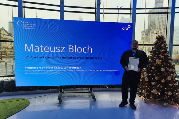 A person stands next to a large blue screen displaying the text: “Mateusz Bloch – First Place in the category for Best Master’s Thesis. Supervisor: Dr Piotr Krzysztof Tronczzyk.” A decorated Christmas tree is visible nearby, with a city skyline and a tall building in the background.