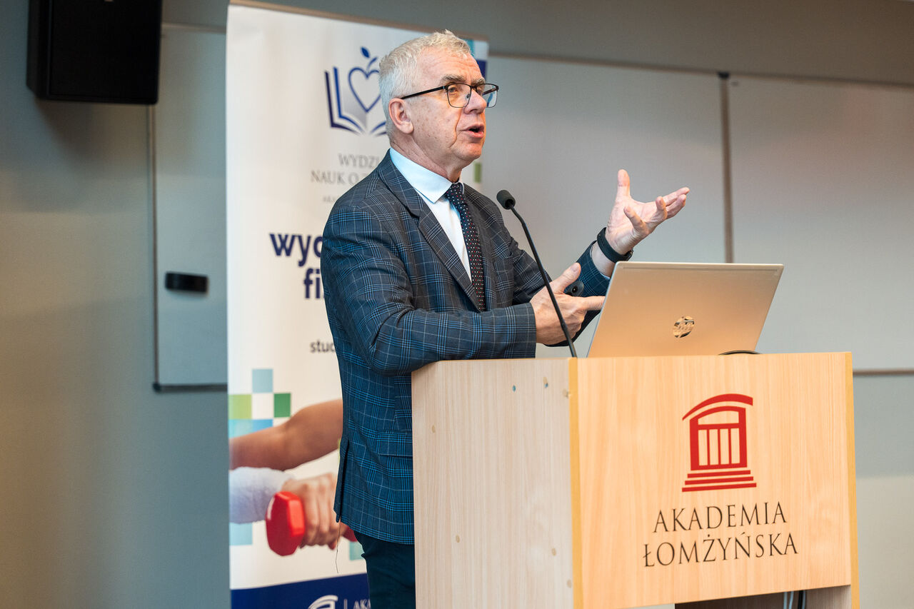A speaker addresses the audience from a lectern bearing the University of Lomza logo, with a banner presenting information about the Faculty of Health Sciences visible nearby.