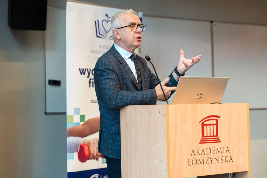 A speaker addresses the audience from a lectern bearing the University of Lomza logo, with a banner presenting information about the Faculty of Health Sciences visible nearby.