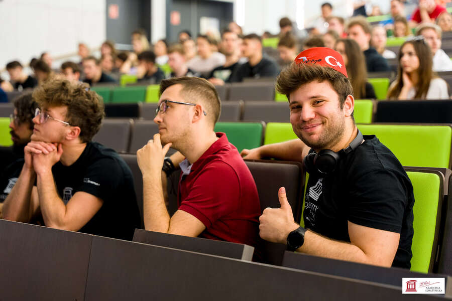 The students are seated in the auditorium, with a student in the foreground showing the "okay" symbol with his thumb raised. He is wearing a traditional Turkish head covering.
