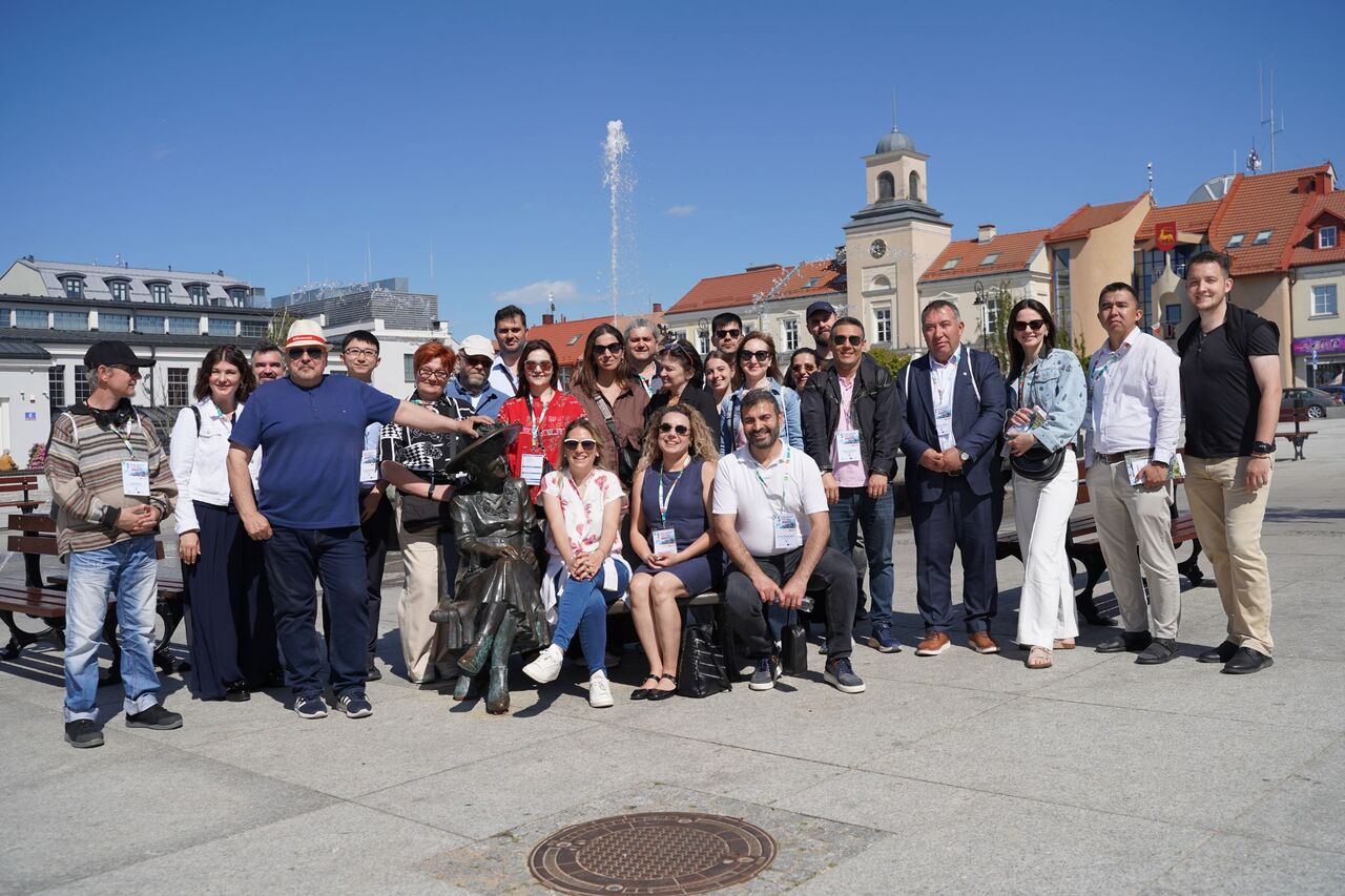 A group of people are posing in the Old Market Square. The town hall can be seen in the background. In the centre of the group is a bench featuring a statue of Hanka Bielicka.
