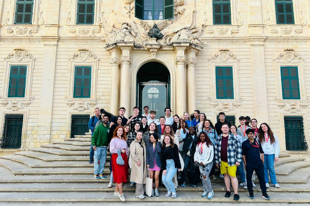 The photo shows a group of students standing in front of the university building.