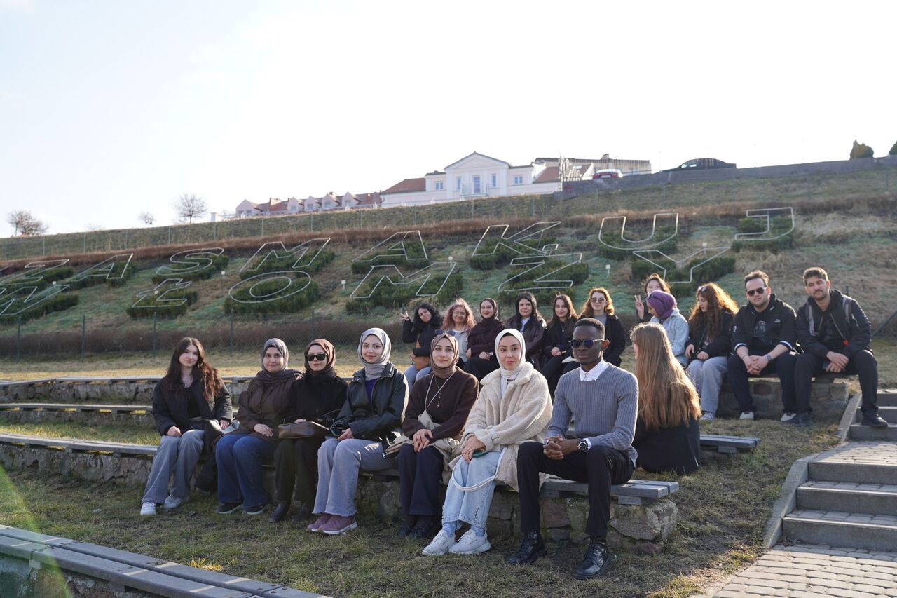Grupa osób pozuje do zdjęcia siedząc na ławkach, na muszli koncertowej. W tle widać napis Zasmakuj w Łomży