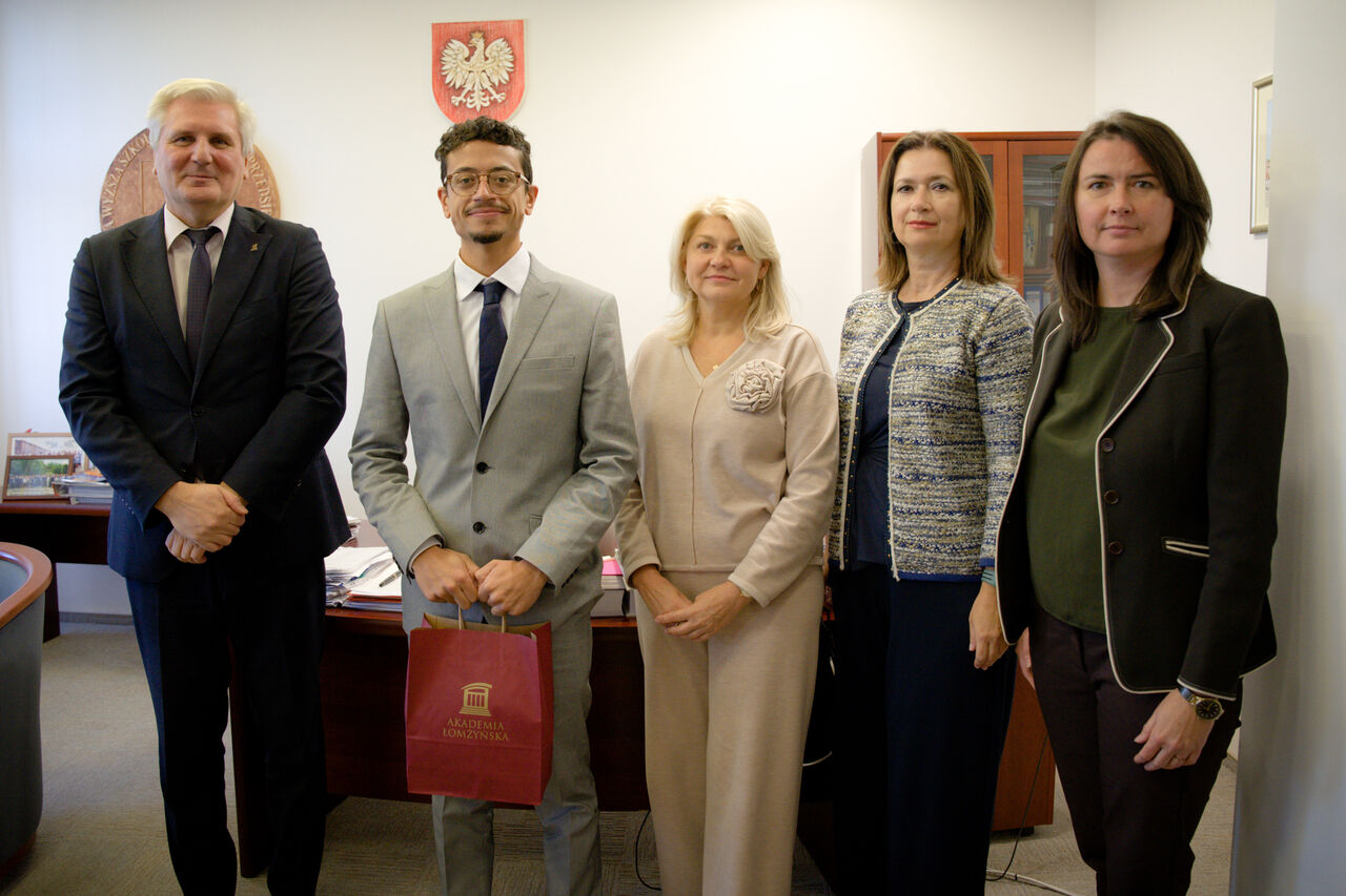 Five people are posing for a photo. On the left, two men are dressed in suits, and on the right, three women are wearing business suits.