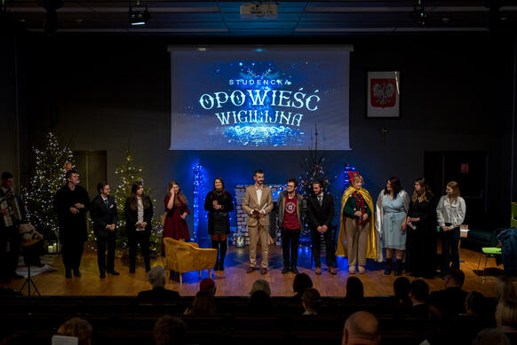 A group of people stands on stage during a performance. Above them, the title “Student Christmas Story” is displayed. The stage is decorated with Christmas trees and festive lights, while a seated audience is visible in front of the stage.