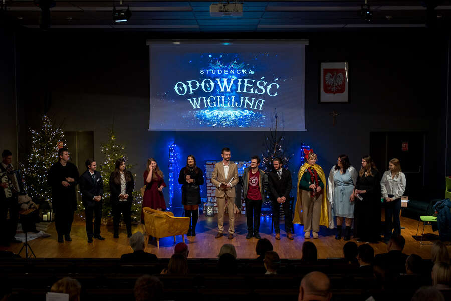 A group of people stands on stage during a performance. Above them, the title “Student Christmas Story” is displayed. The stage is decorated with Christmas trees and festive lights, while a seated audience is visible in front of the stage.