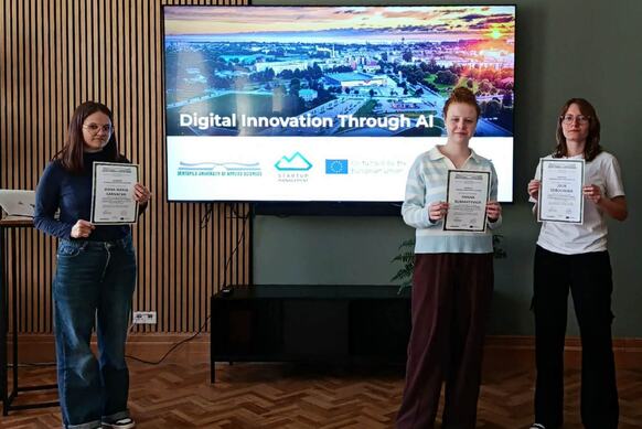 Three female students stand in front of a screen, holding certificates in their hands
