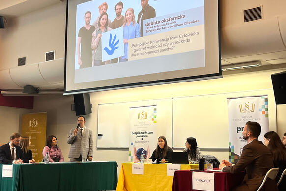 An Oxford-style debate in a lecture hall, with three tables of participants, organisers’ banners, and a screen displaying a presentation on the European Convention on Human Rights.