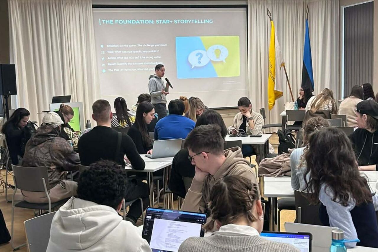 A room full of students seated at small tables, with a presentation and a speaker visible in the background.