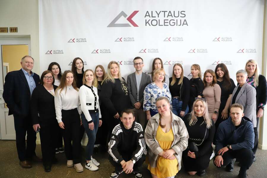 Studenci i wykładowcy na tle ściany z logo uczelni