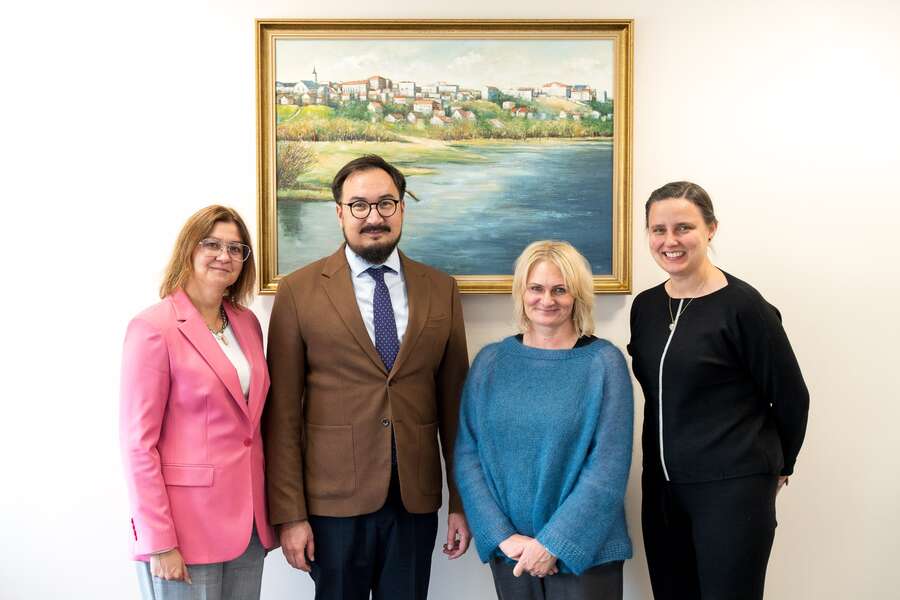 Four people posing for a photo: three women and a man positioned in the center. All are dressed elegantly in blazers and suits. A painting is visible in the background
