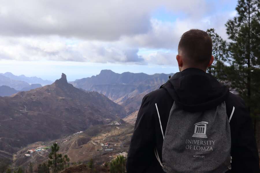 Mężczyzna z plecakiem na plecach na którym widać logo University of Lomza, patrzy w stronę gór