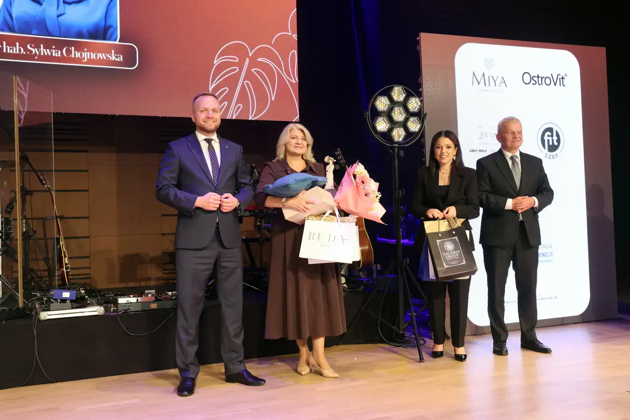 „Cztery elegancko ubrane osoby stoją na scenie podczas uroczystości. Kobieta w środku trzyma kwiaty i prezenty, pozostali również mają torby upominkowe. W tle widać instrumenty muzyczne oraz ekrany z logotypami sponsorów.”