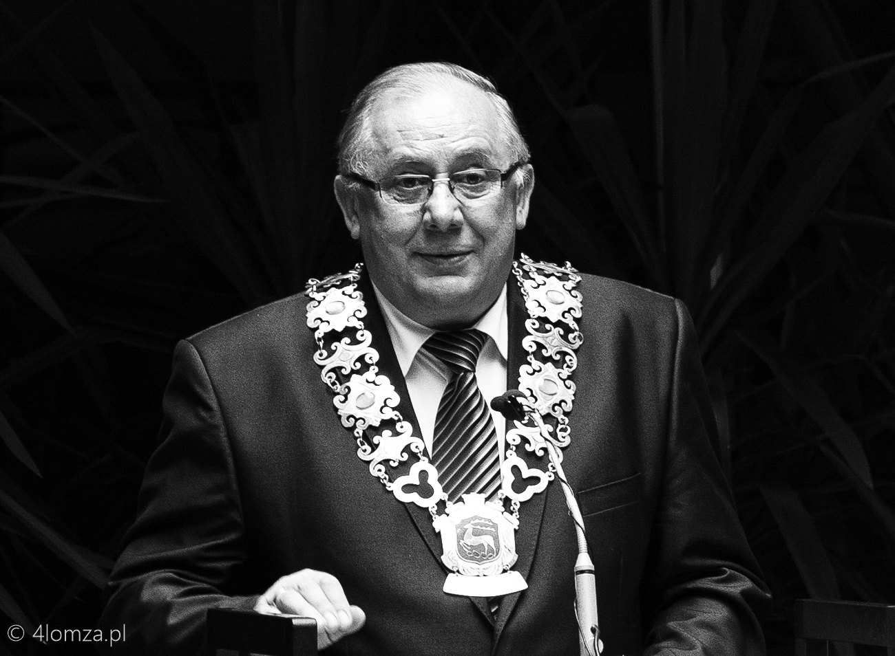 Mężczyzna w garniturze z łańcuchem urzędowym stoi przy mównicy i przemawia.