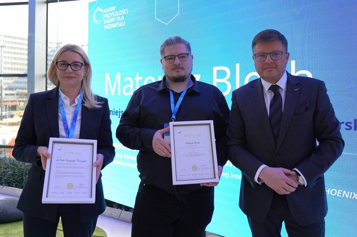 Trzy osoby stoją przed dużym niebieskim ekranem z napisem ‘Mateusz Bloch’. Dwie osoby trzymają dyplomy w jasnych ramkach, a trzecia osoba stoi obok w eleganckim stroju. W tle widoczny fragment napisu i nowoczesne wnętrze z dużymi oknami.