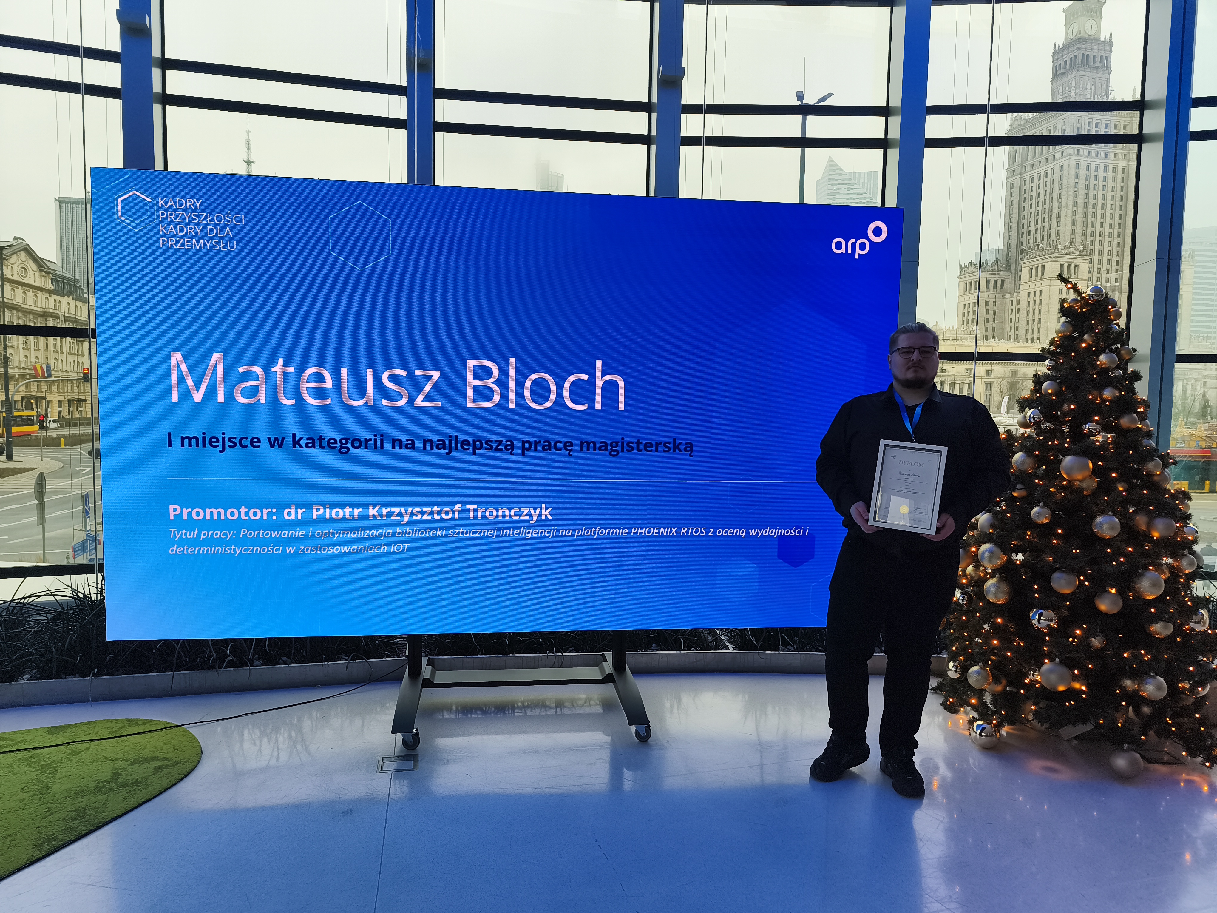 Osoba stojąca obok dużego niebieskiego ekranu z napisem: ‘Mateusz Bloch – I miejsce w kategorii na najlepszą pracę magisterską. Promotor: dr Piotr Krzysztof Tronczzyk’. Obok znajduje się udekorowana choinka, w tle widać panoramę miasta z wysokim budynkiem.