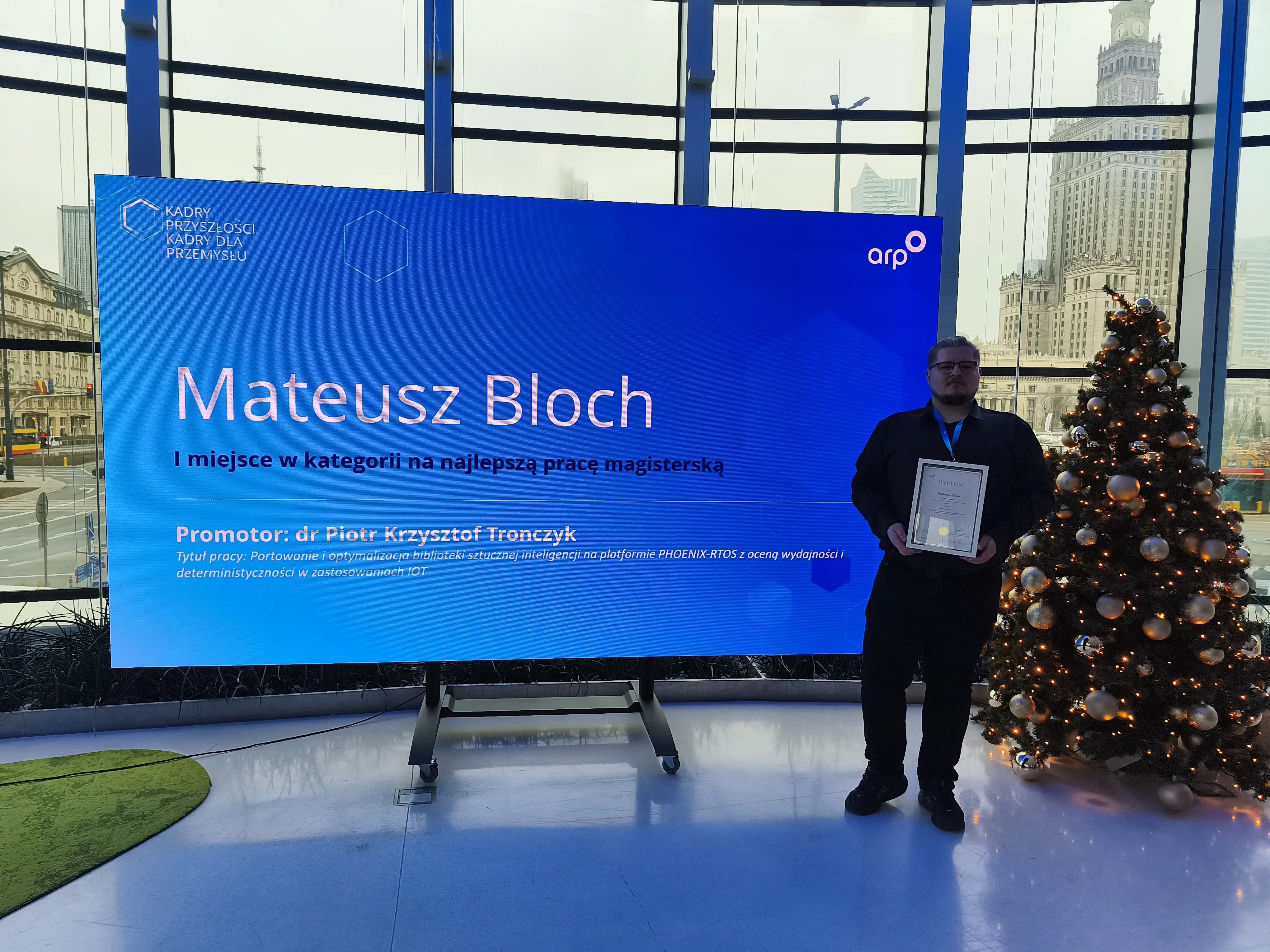 Osoba stojąca obok dużego niebieskiego ekranu z napisem: ‘Mateusz Bloch – I miejsce w kategorii na najlepszą pracę magisterską. Promotor: dr Piotr Krzysztof Tronczzyk’. Obok znajduje się udekorowana choinka, w tle widać panoramę miasta z wysokim budynkiem.