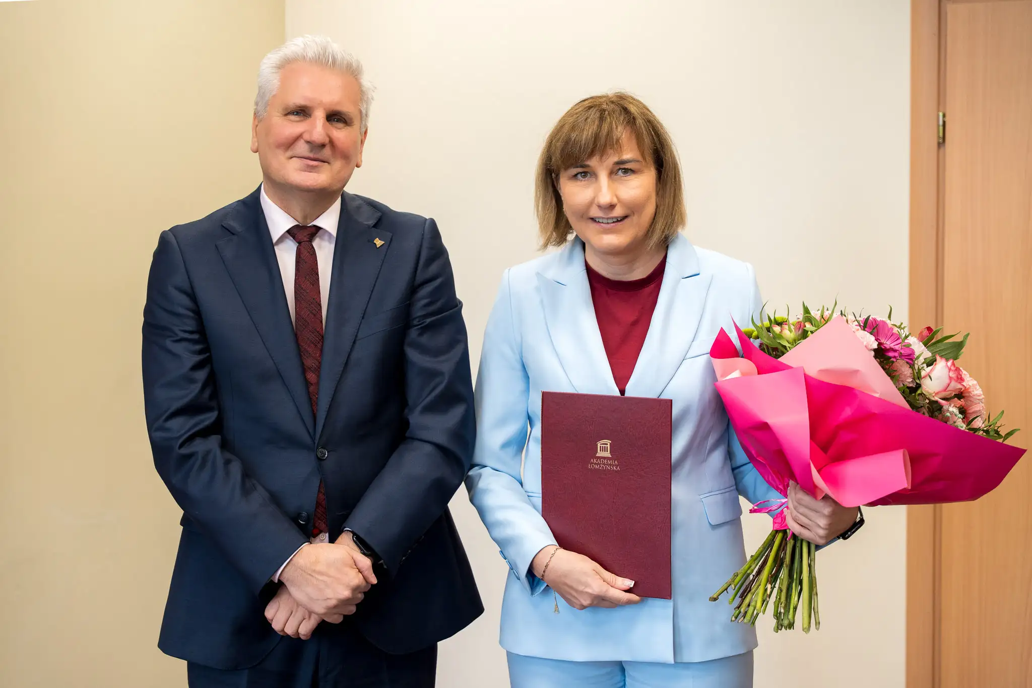 dwie osoby pozują do zdjęcia, kobieta trzyma teczkę i kwiaty