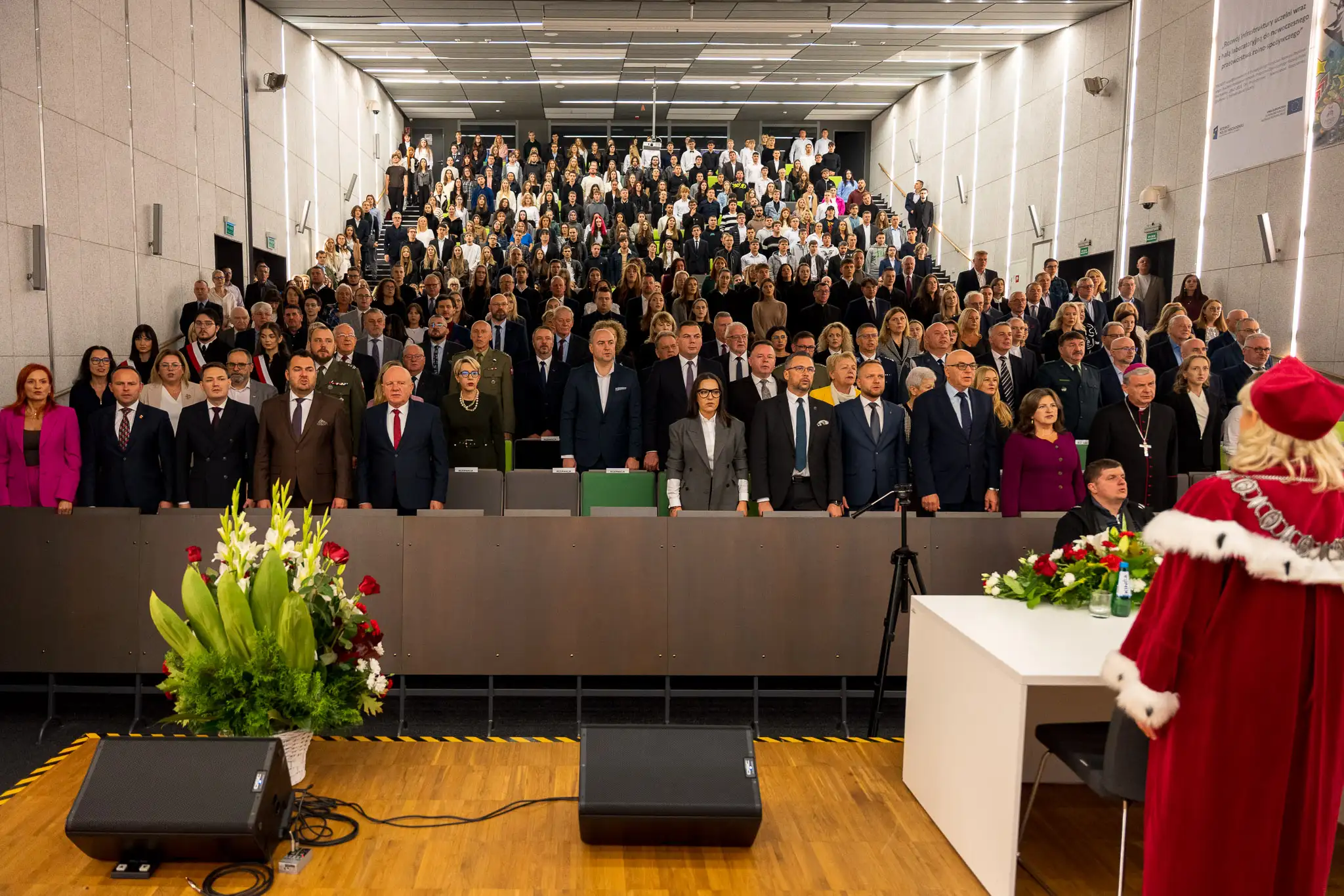 Duża sala audytoryjna wypełniona ludźmi stojącymi podczas uroczystej akademickiej ceremonii, z przodu osoba w czerwonym todze zwrócona plecami.
