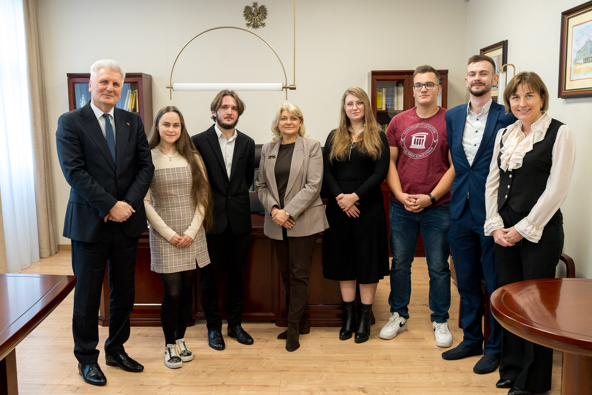 Osiem osób pozuje do wspólnego zdjęcia w biurze. W tle widać biurko, szafki z książkami i godło na ścianie.