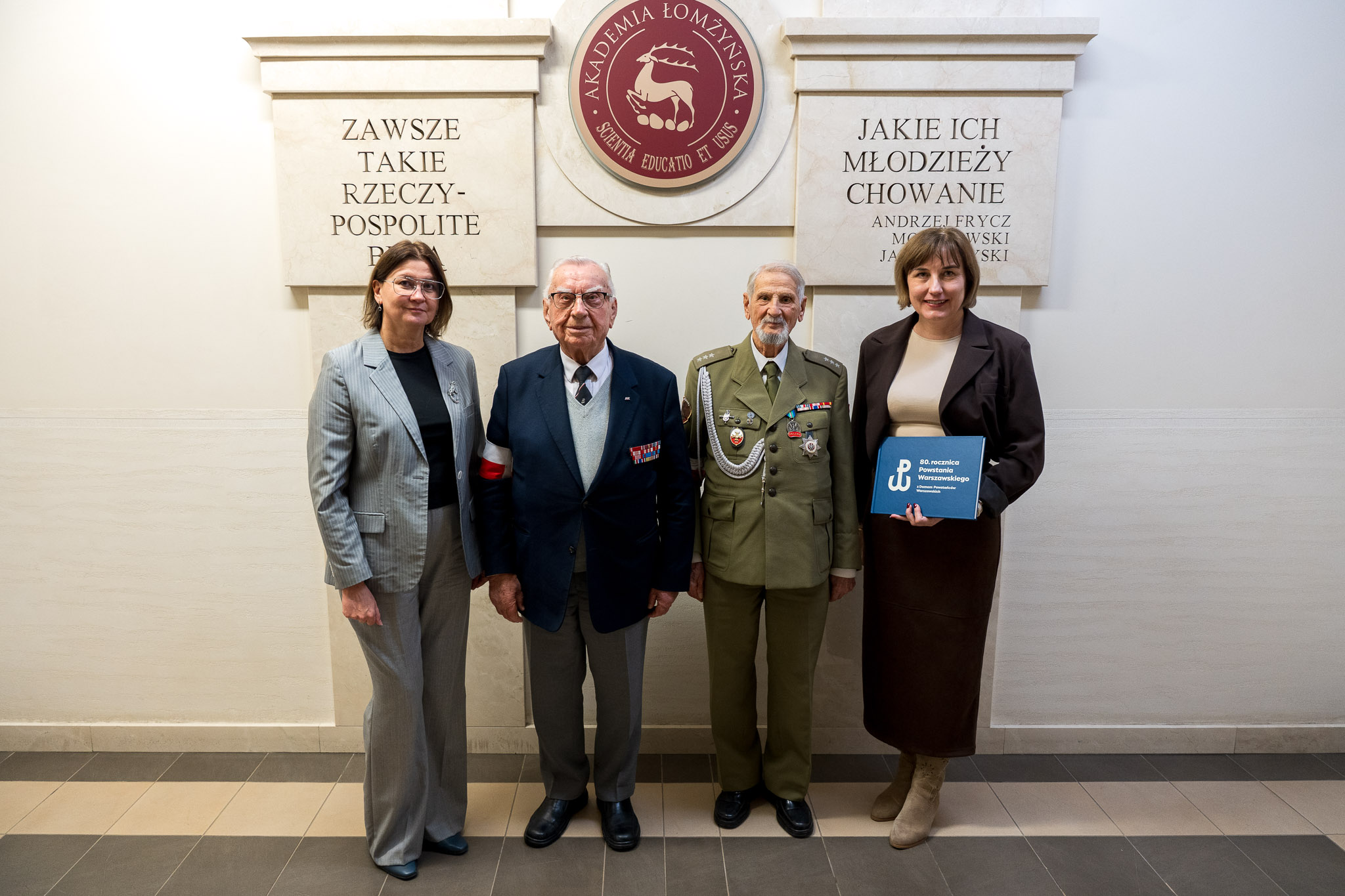 Cztery osoby stoją przed tablicą z napisem Akademia Łomżyńska, w tym dwie w strojach wojskowych, jedna trzyma niebieską teczkę.