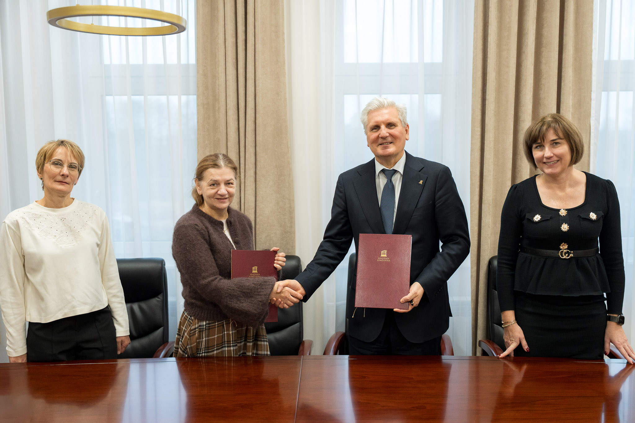 Cztery osoby stoją przy stole konferencyjnym, dwie z nich trzymają teczki i podają sobie ręce.