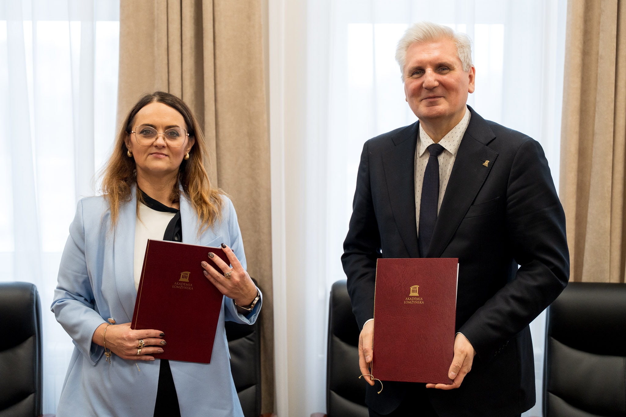 Dwie osoby stoją obok siebie, trzymając teczki z logo uczelni.