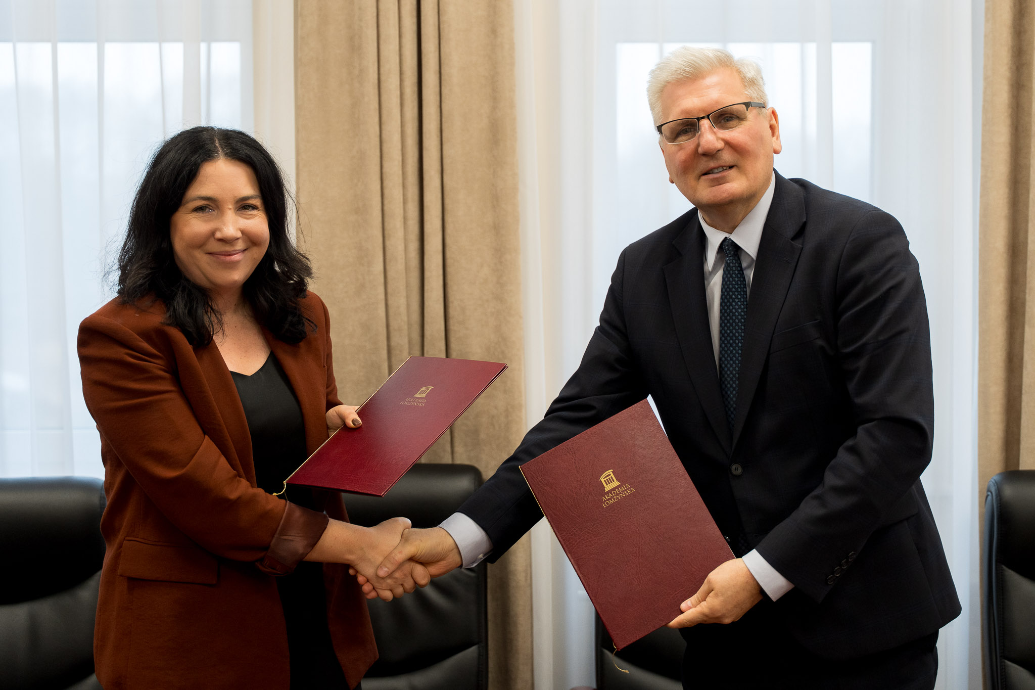 Dwie osoby stojące w sali konferencyjnej, podają sobie ręce i trzymają bordowe teczki z logo instytucji.