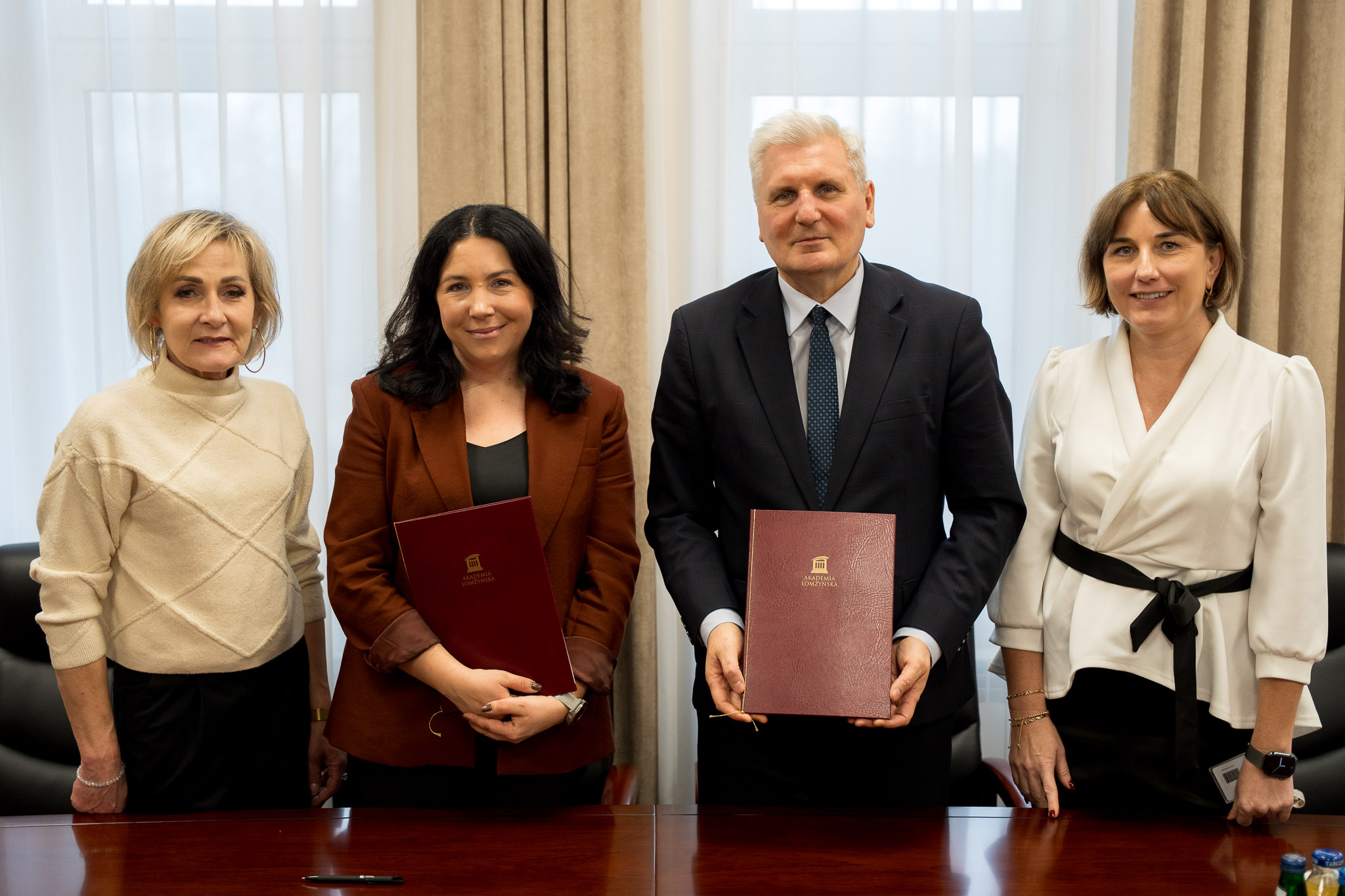 Cztery osoby stojące przy stole w sali konferencyjnej, dwie z nich trzymają bordowe teczki z logo instytucji