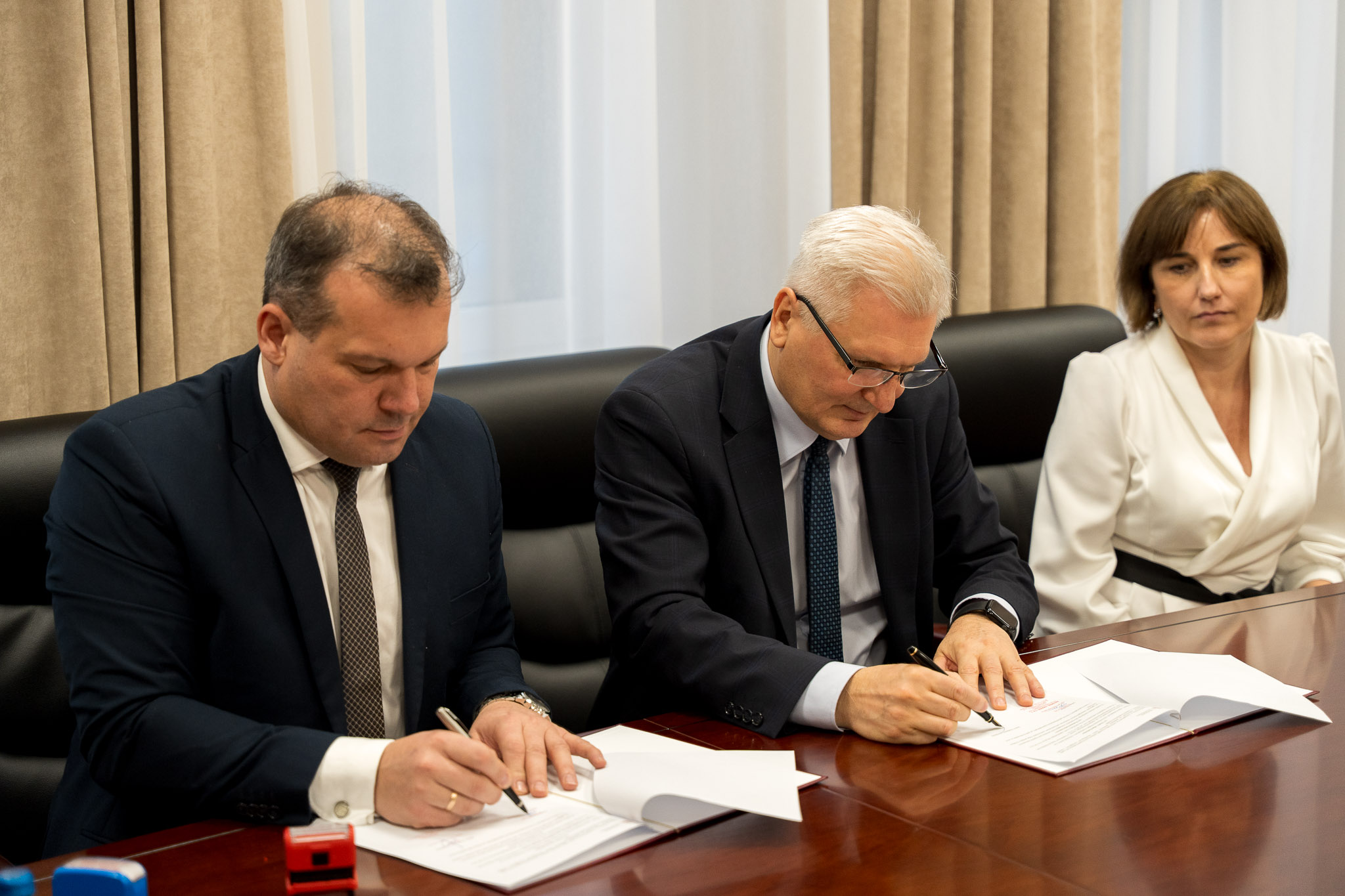 Dwie osoby siedzą przy stole i podpisują dokumenty, obok nich siedzi trzecia osoba.