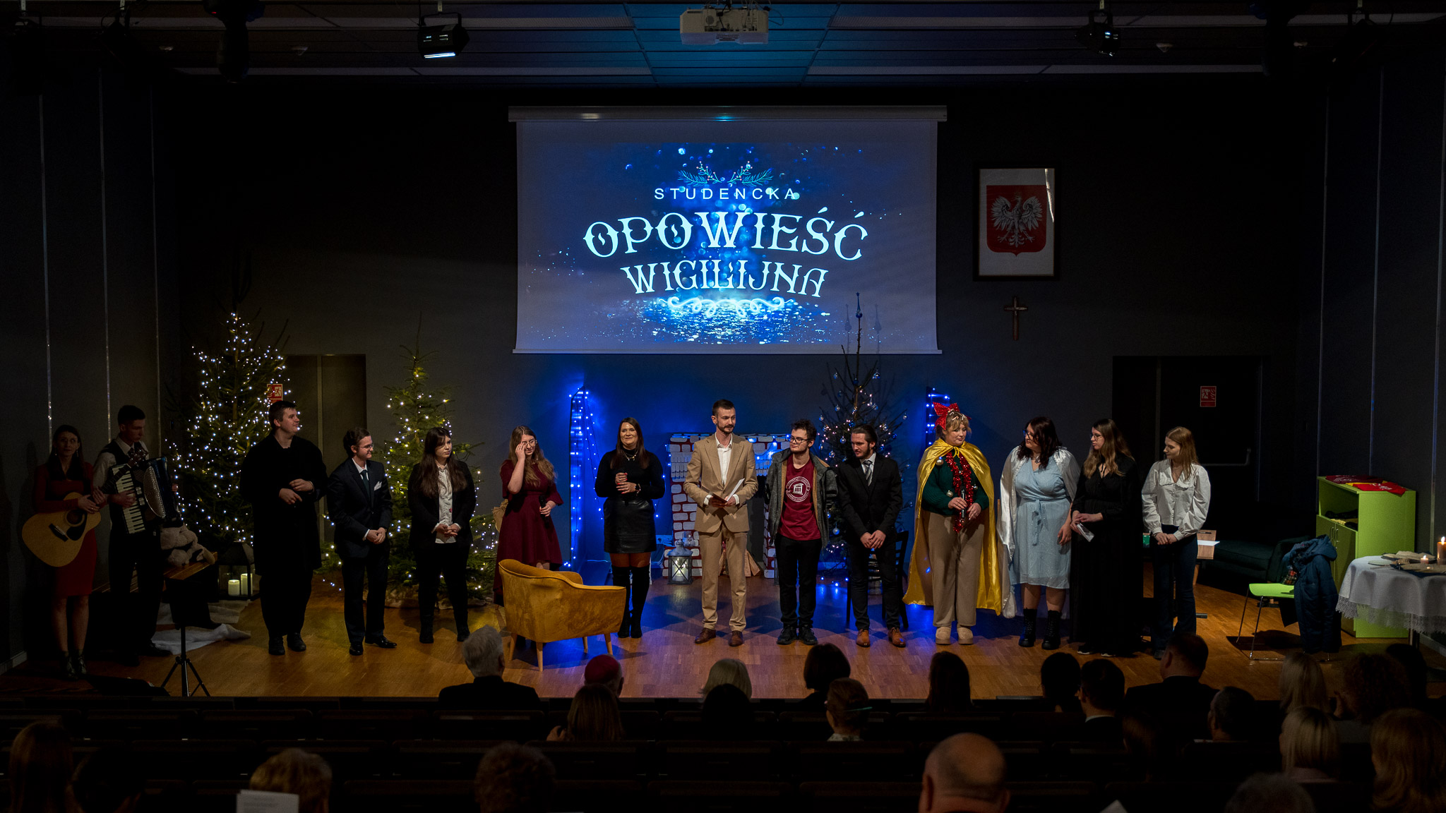 Grupa osób stoi na scenie podczas przedstawienia. Nad nimi wyświetlany jest napis „Studencka Opowieść Wigilijna”. Scena jest udekorowana choinkami i świątecznymi lampkami. Przed sceną widoczna jest siedząca publiczność.
