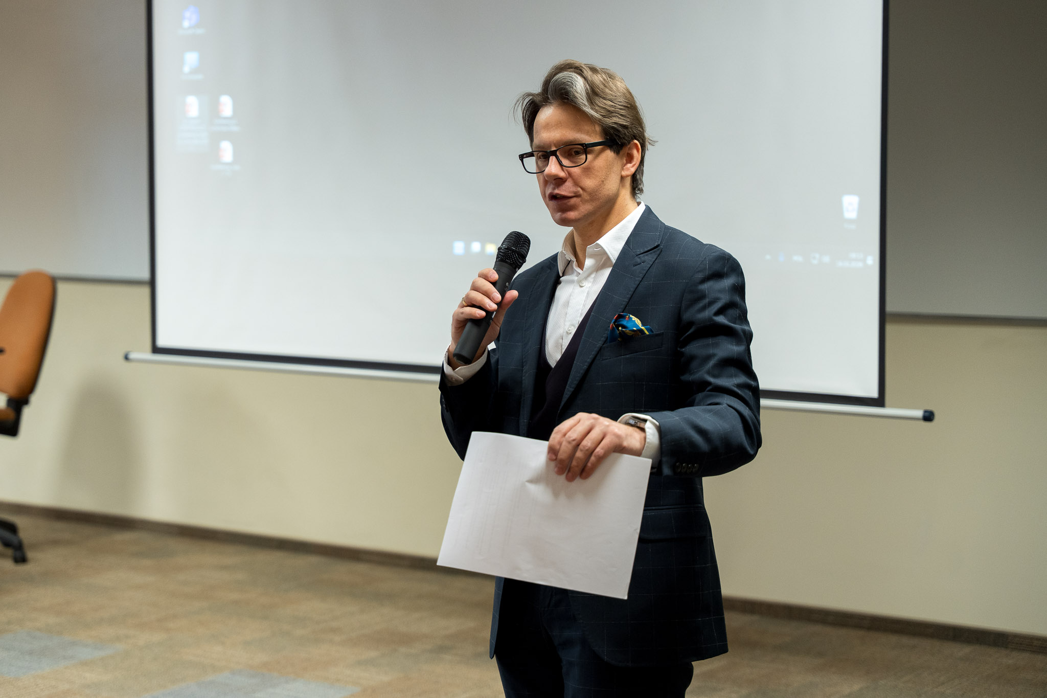 Osoba w garniturze przemawia przed ekranem projekcyjnym w sali konferencyjnej