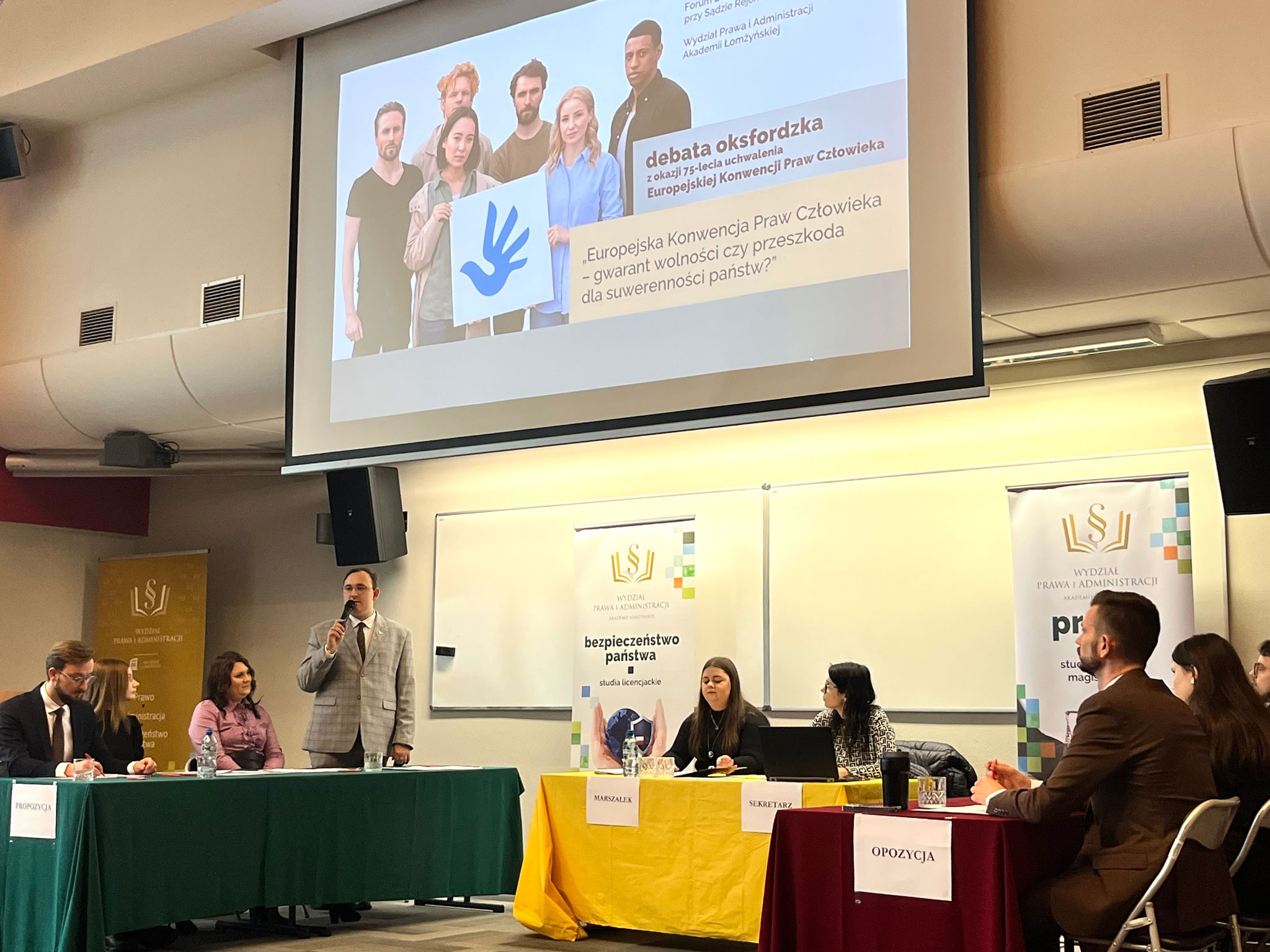 Debata oksfordzka w sali wykładowej – trzy stoły z uczestnikami, banery organizatora, ekran z prezentacją o Europejskiej Konwencji Praw Człowieka.
