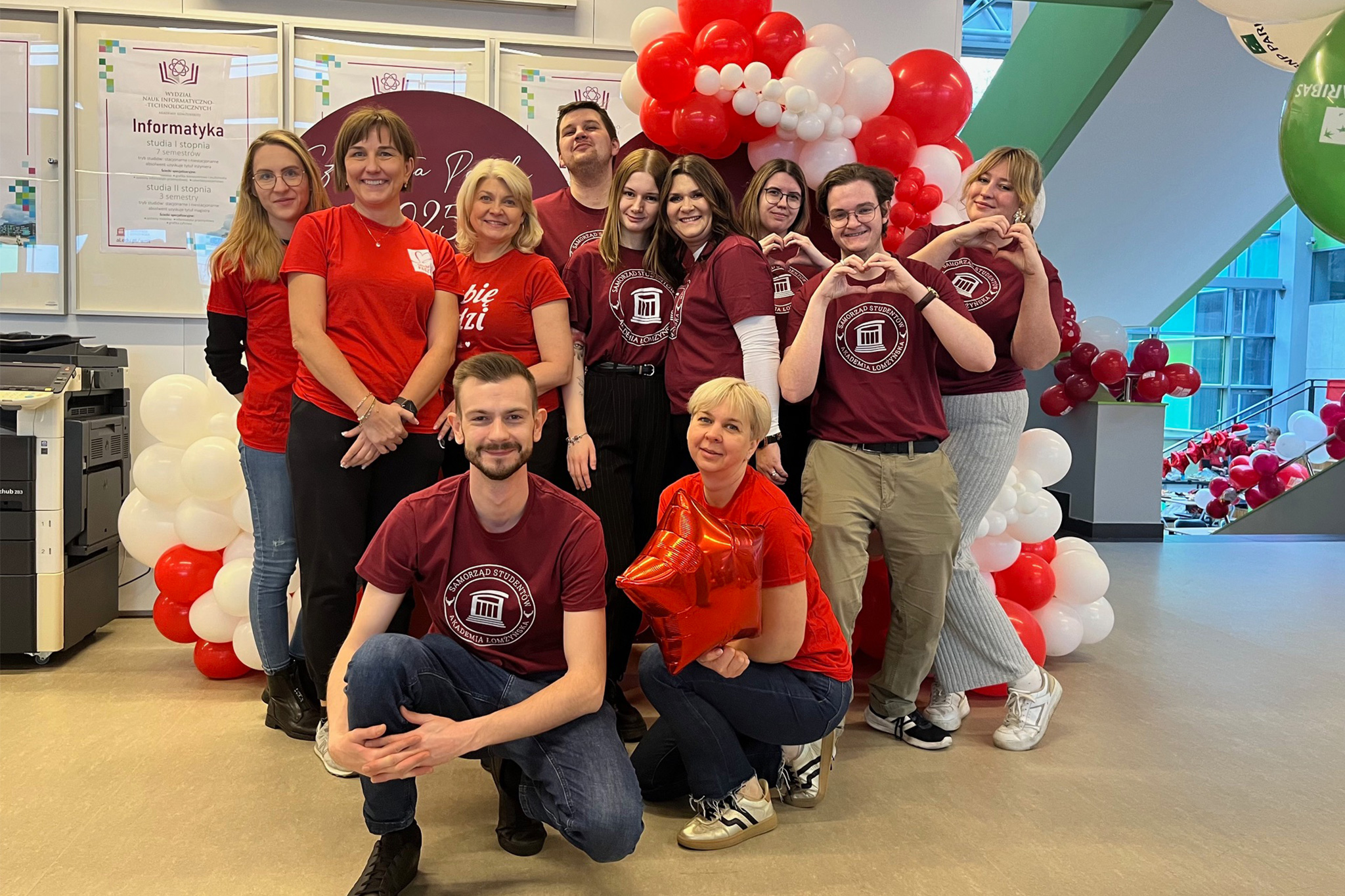 Grupa osób stoi i kuca przed dekoracją z czerwonych i białych balonów w pomieszczeniu. Większość osób ma na sobie bordowe koszulki z białym nadrukiem, a dwie osoby noszą czerwone koszulki. W tle widoczne są plakaty informacyjne oraz balony w różnych kolorach, w tym zielony po prawej stronie.