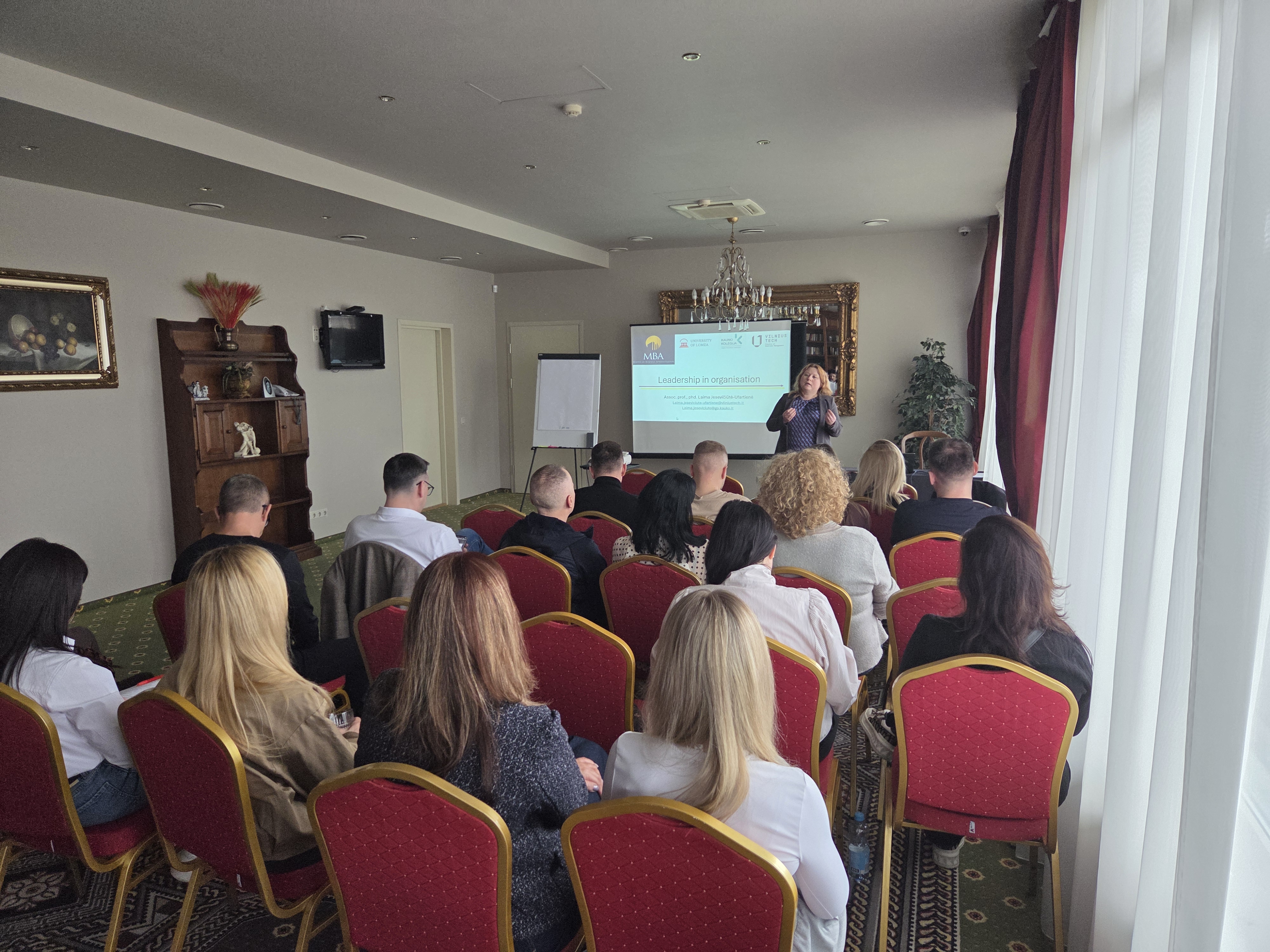 Uczestnicy siedzą na sali konferencyjnej i słuchają prezentacji prowadzonej przed ekranem projekcyjnym