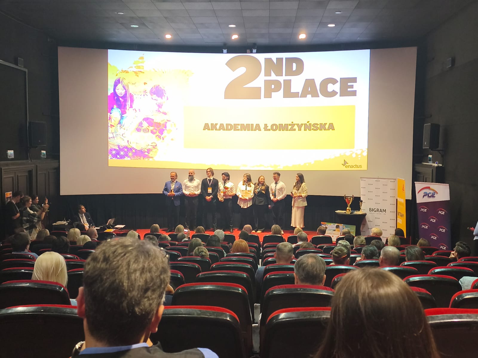 Uczestnicy stoją na scenie podczas wydarzenia, a na ekranie wyświetlony jest napis ‘2nd Place Akademia Łomżyńska’, przed publicznością w sali.