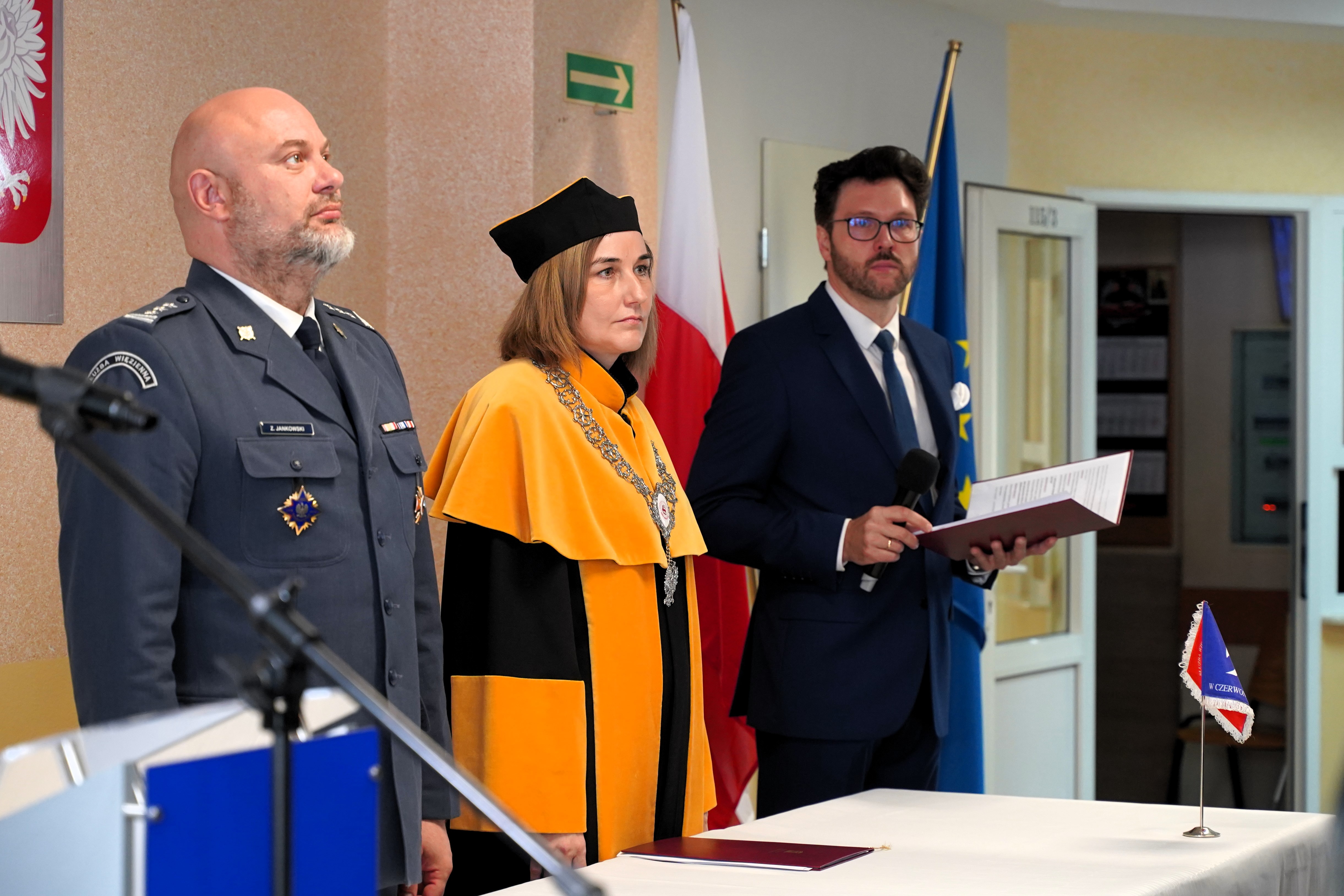 Trzy osoby stoją przy stole podczas uroczystości, na tle flag Polski i Unii Europejskiej oraz symboli państwowych